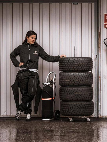 Frau in CUPRA Outfit lehnt an einem Stapel Autoreifen neben einem CUPRA Rennfahrzeug in einer Garage.